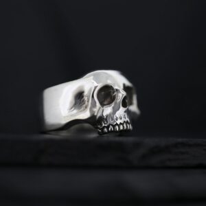 A silver skull ring with detailed eye sockets and teeth on a dark background.