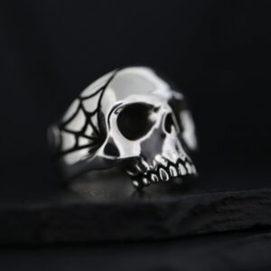 A silver skull ring with spider web details on a dark background.