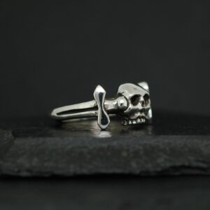 Silver ring featuring a skull and dagger design on a dark background.