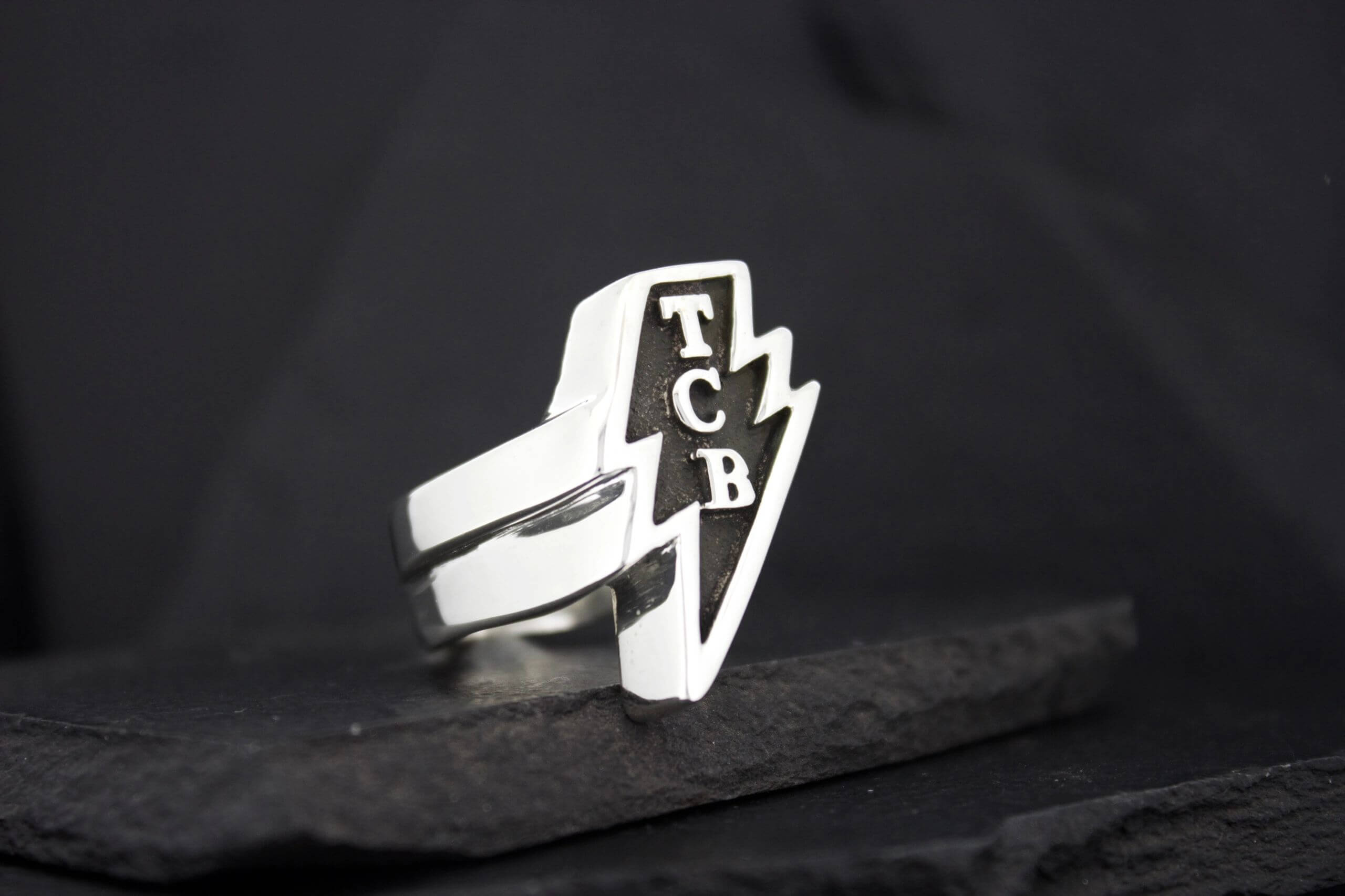 A silver ring with a lightning bolt and 'TCB' initials.