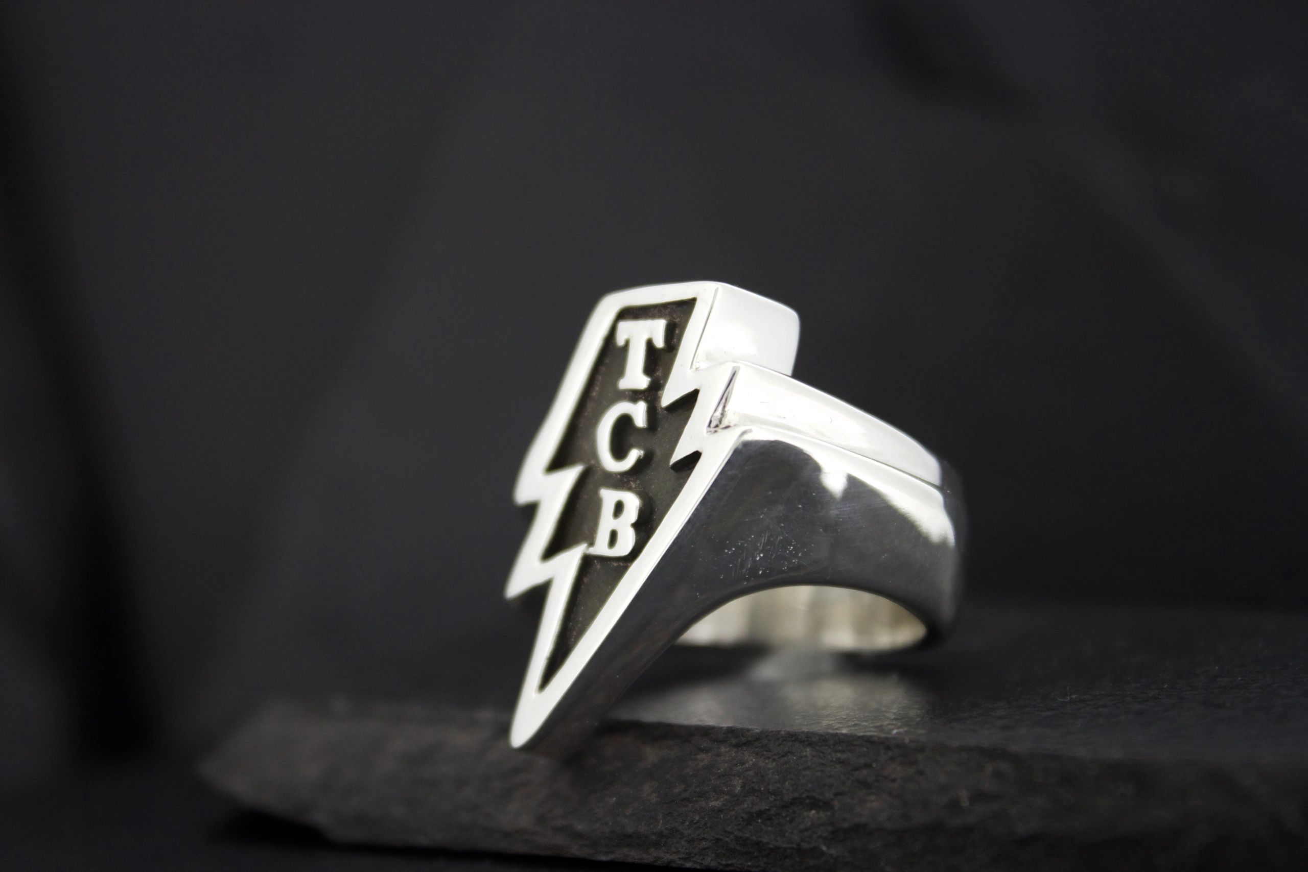 Silver ring with TCB lightning bolt design on a black background.