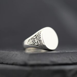 Silver signet ring with intricate side engravings on a dark surface.