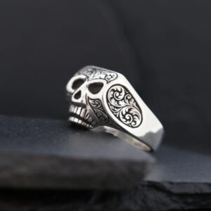 A silver skull ring with intricate engravings on a dark background.