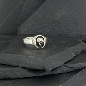 Silver ring with a skull design resting on dark slate stones.