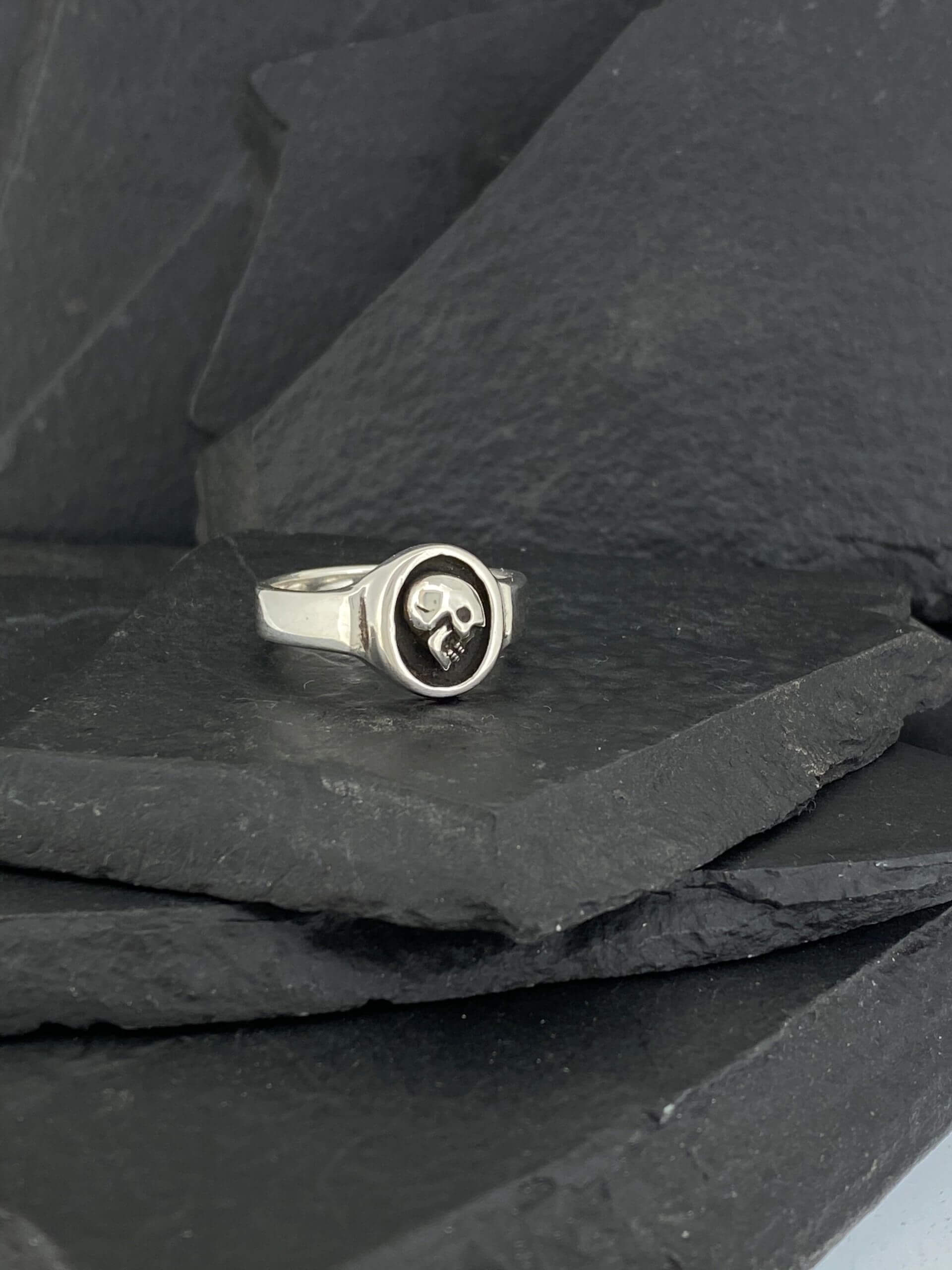 Silver ring with a skull design resting on dark slate stones.