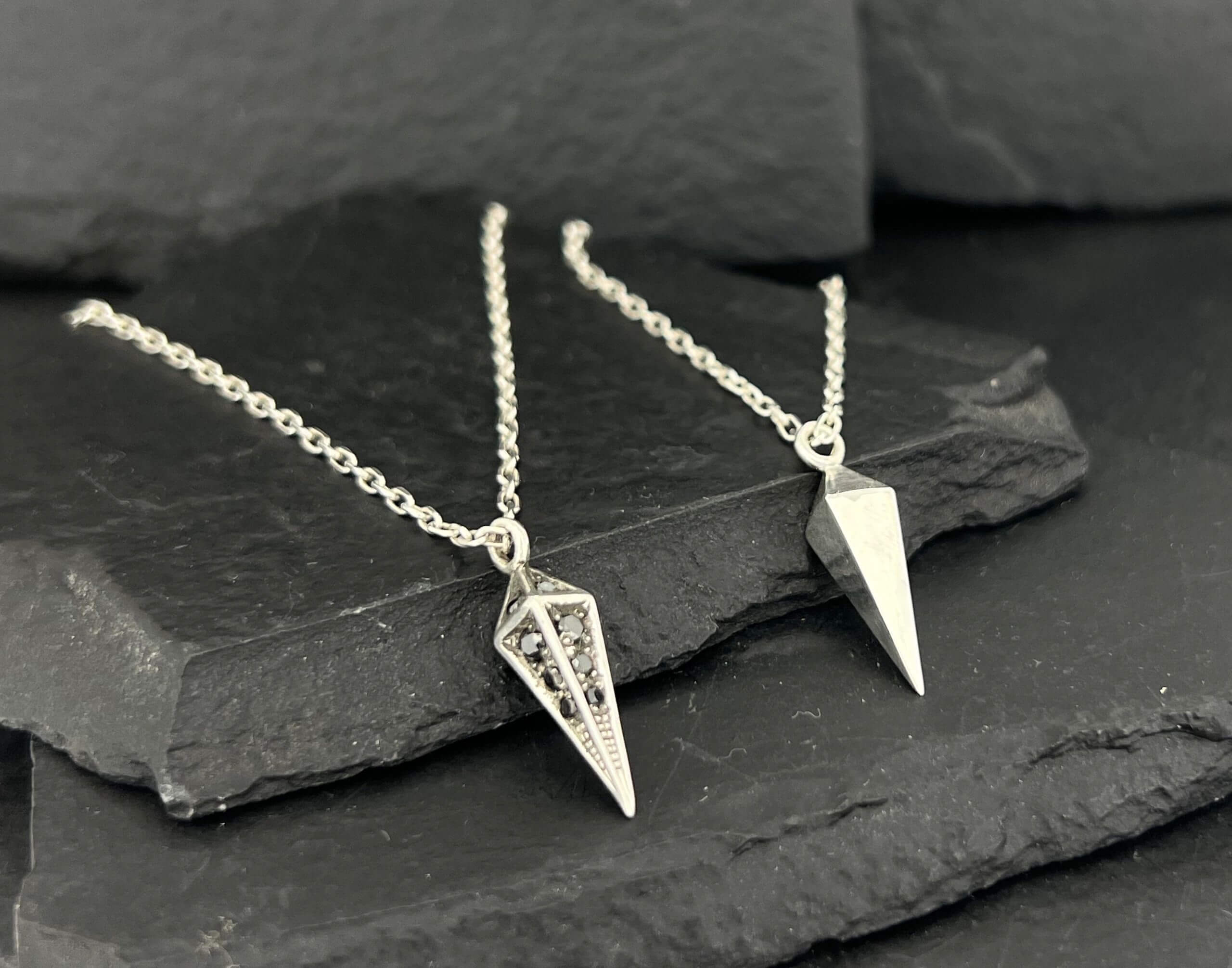Two silver geometric pendant necklaces on a dark stone surface.