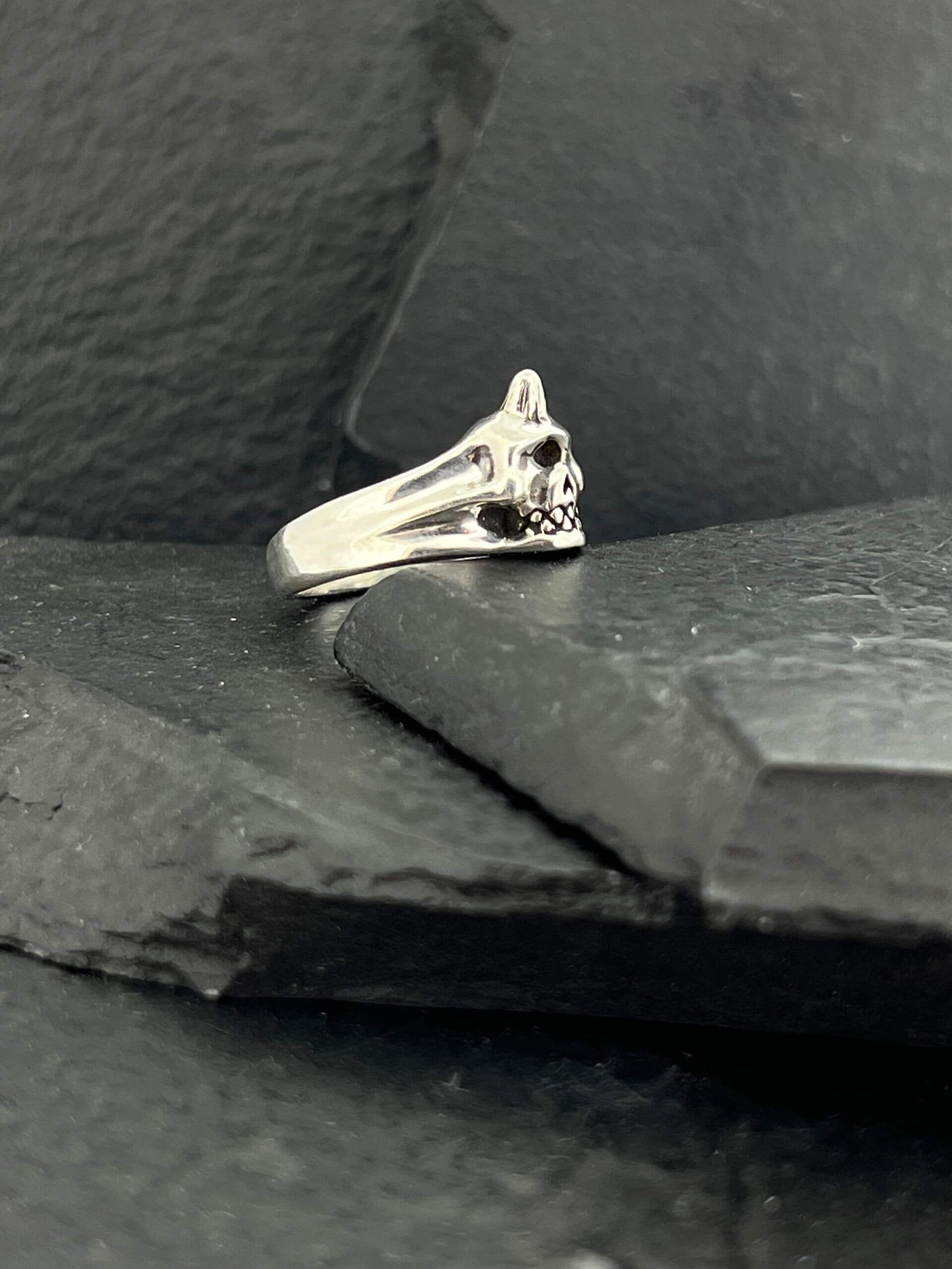 Silver ring with a skull design on a dark textured surface.