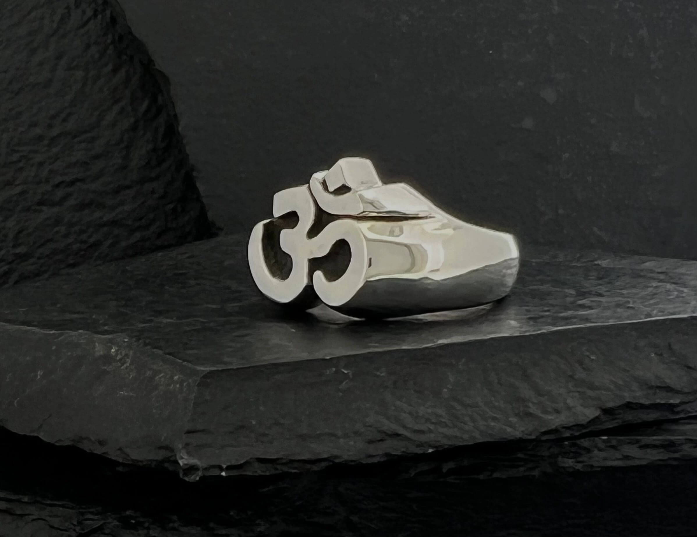 Silver ring featuring an Om symbol design on a black textured surface.