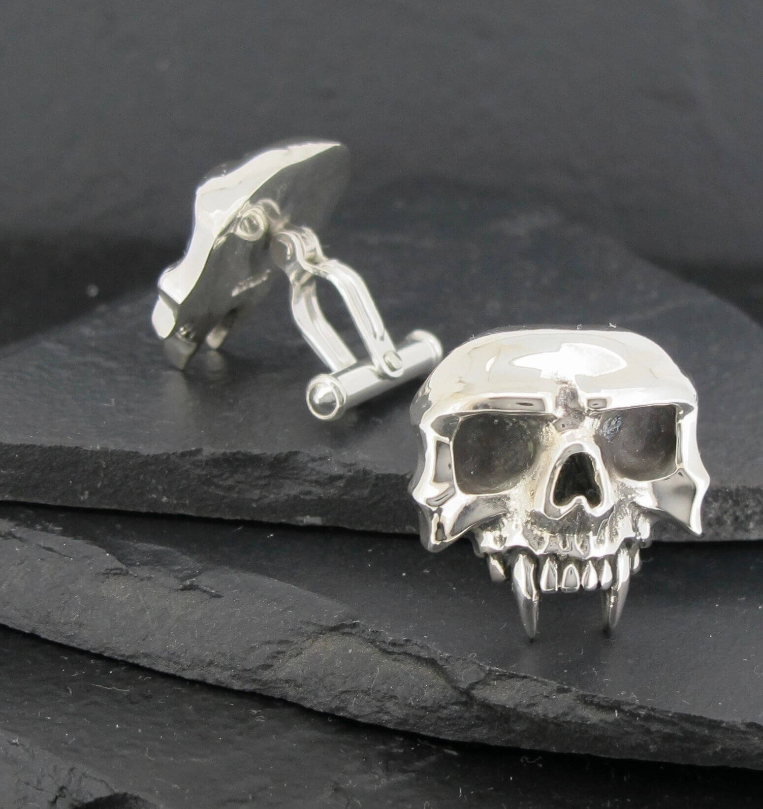 Silver skull cufflinks with detailed fangs on a dark slate surface.