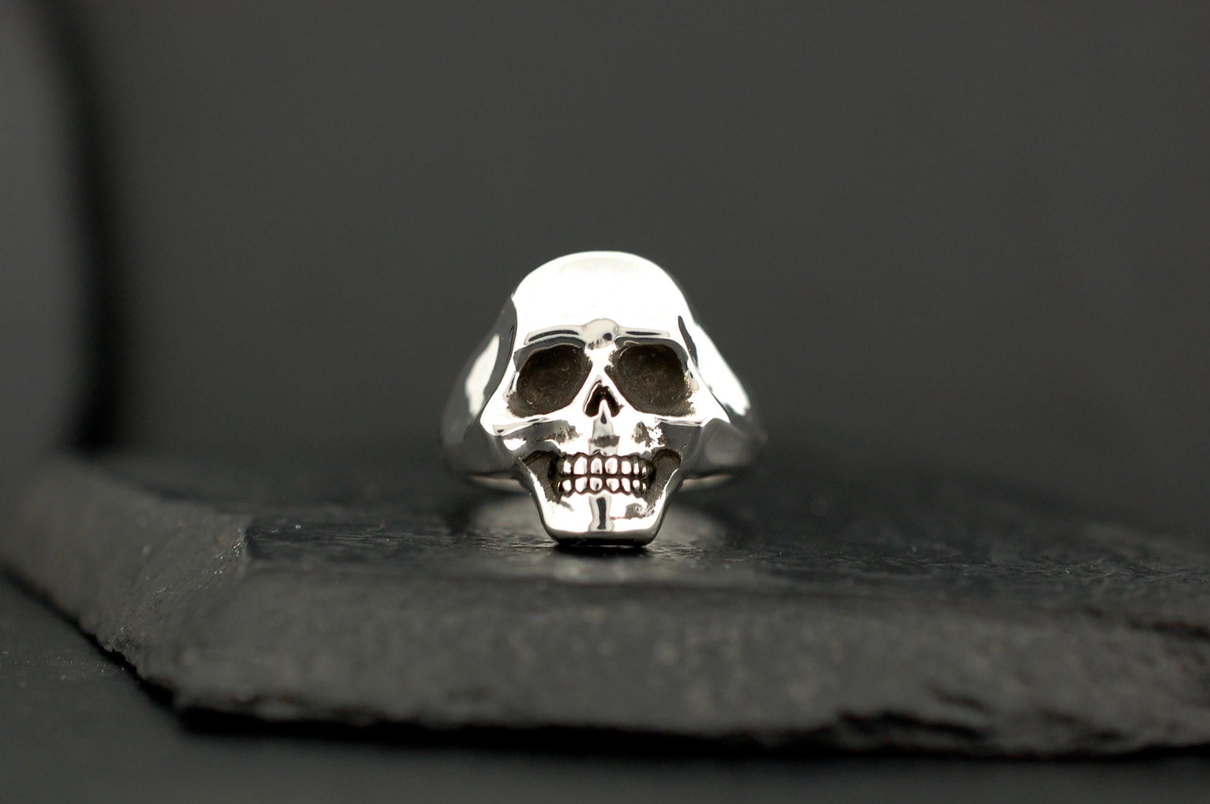 A silver skull ring with detailed facial features on a dark surface.