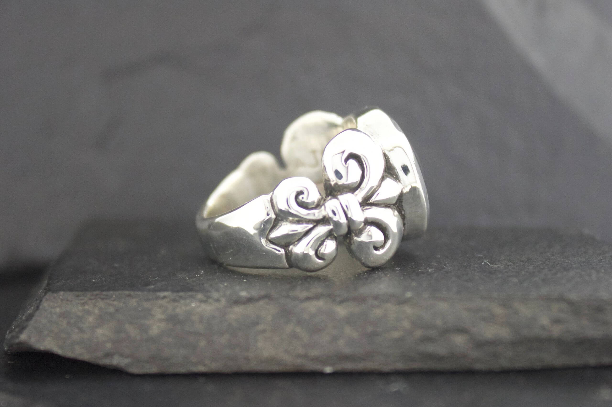 Silver ring with floral and swirl design on a dark stone surface.