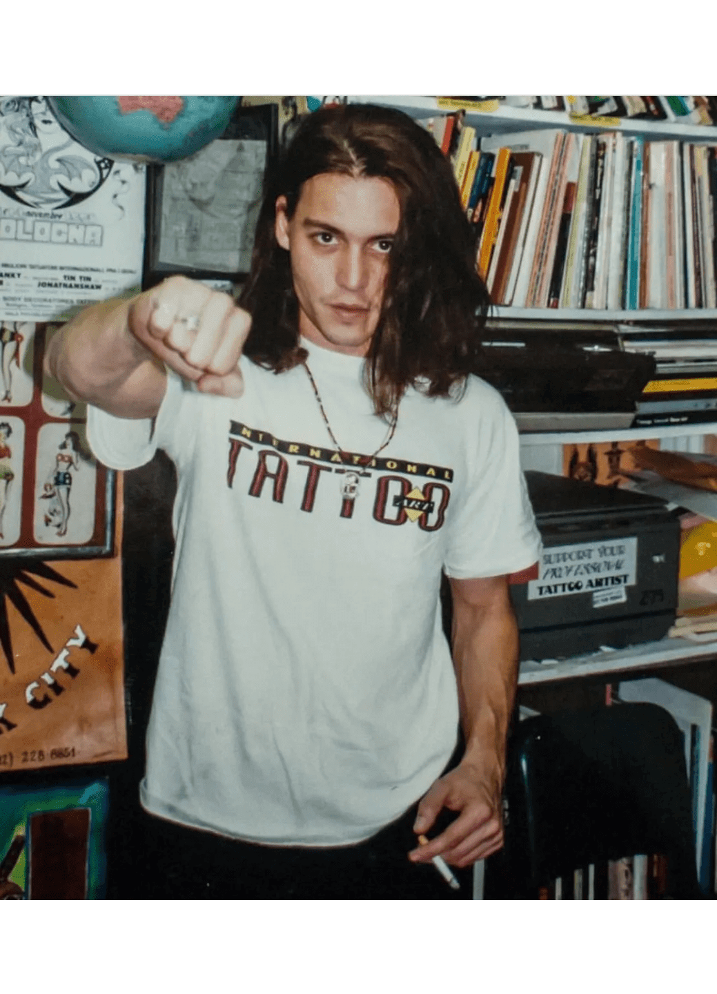 Young man with long hair pointing, wearing a white T-shirt with 'TATTOO' text.